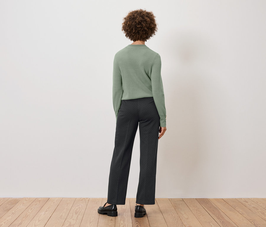 Rückansicht einer Frau mit lockigem Haar, die einen grünen Pullover und dunkle Hosen trägt und auf einem Holzboden vor einer weißen Wand steht.