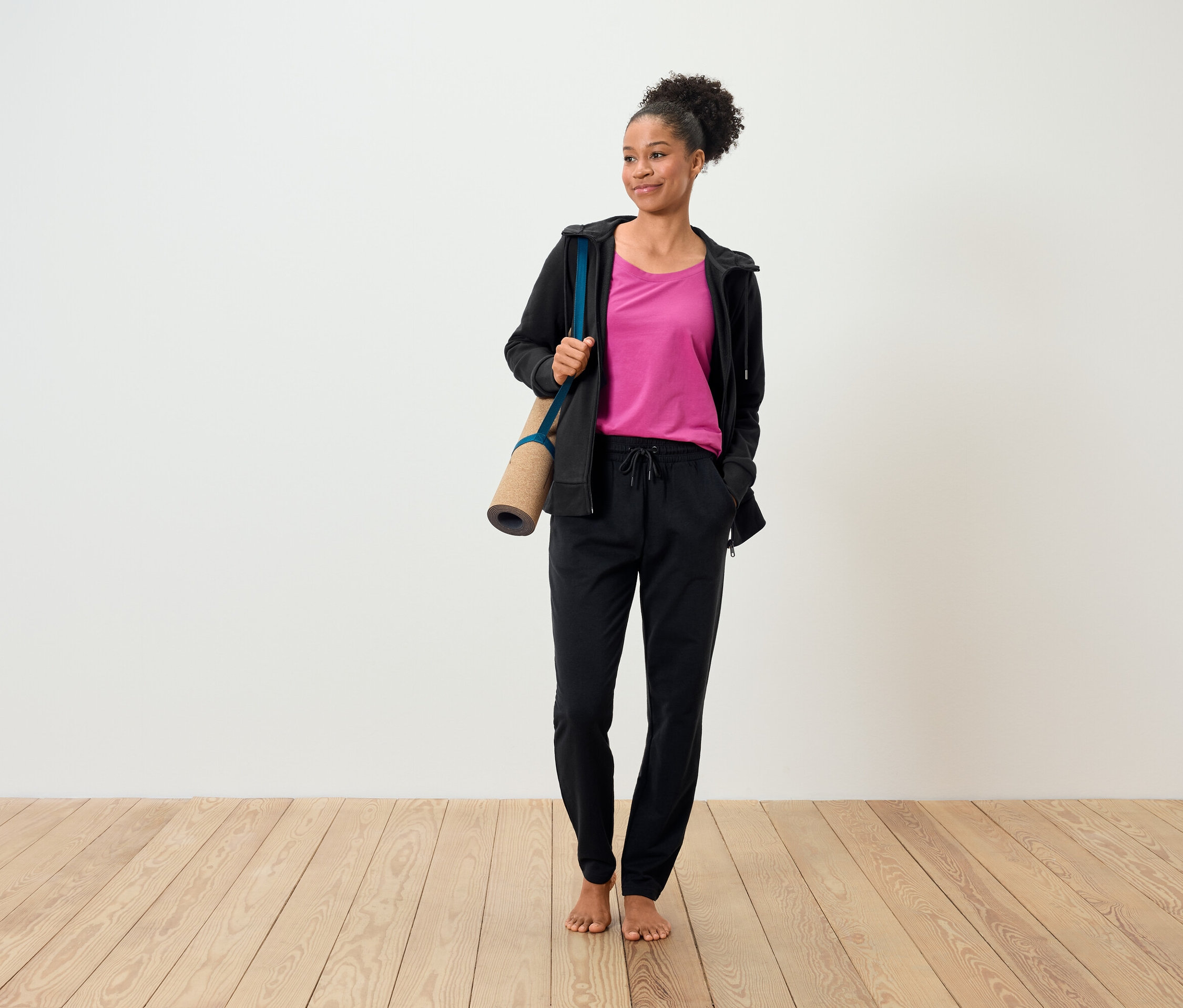 Frau mit braunen Haaren hält eine Yogamatte und trägt ein rosa T-Shirt, eine schwarze Sweatjacke und eine schwarze Hose.