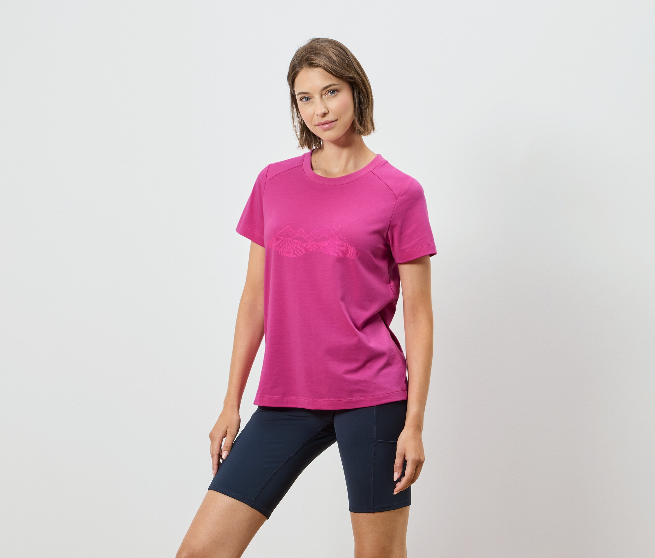Frau trägt ein pinkes T-Shirt mit einer Bergkette und dunkelblaue Shorts vor einem weißen Hintergrund.