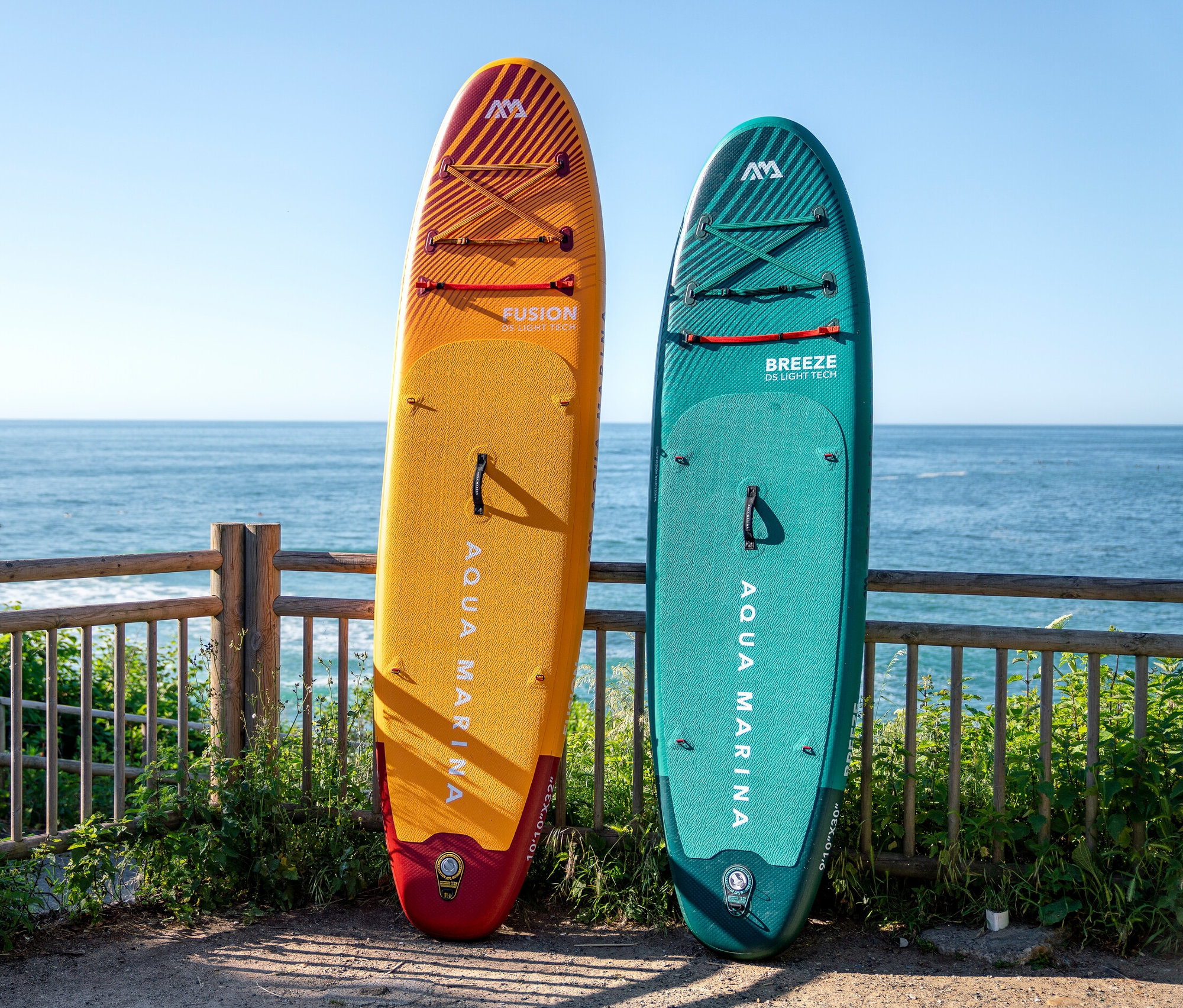 Gelbes und grünes Aqua Marina SUP Board »Fusion« 10'10" lehnt an einem Holzzaun mit Blick auf den Ozean.