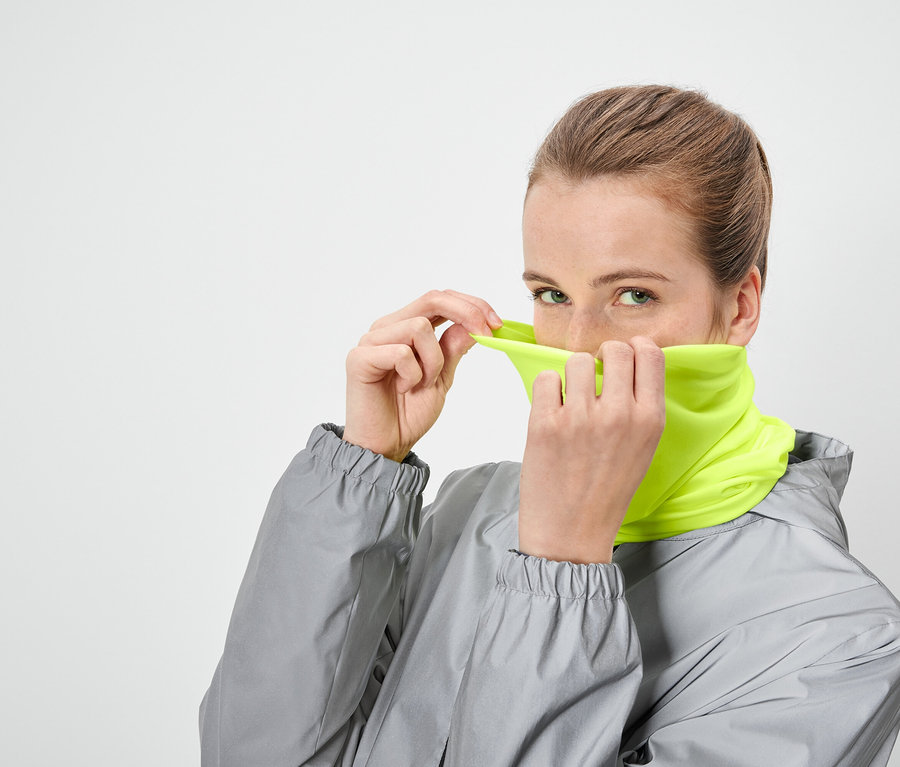Frau zieht ein neongelbes Schlauchtuch über ihr Gesicht und trägt eine graue Jacke.