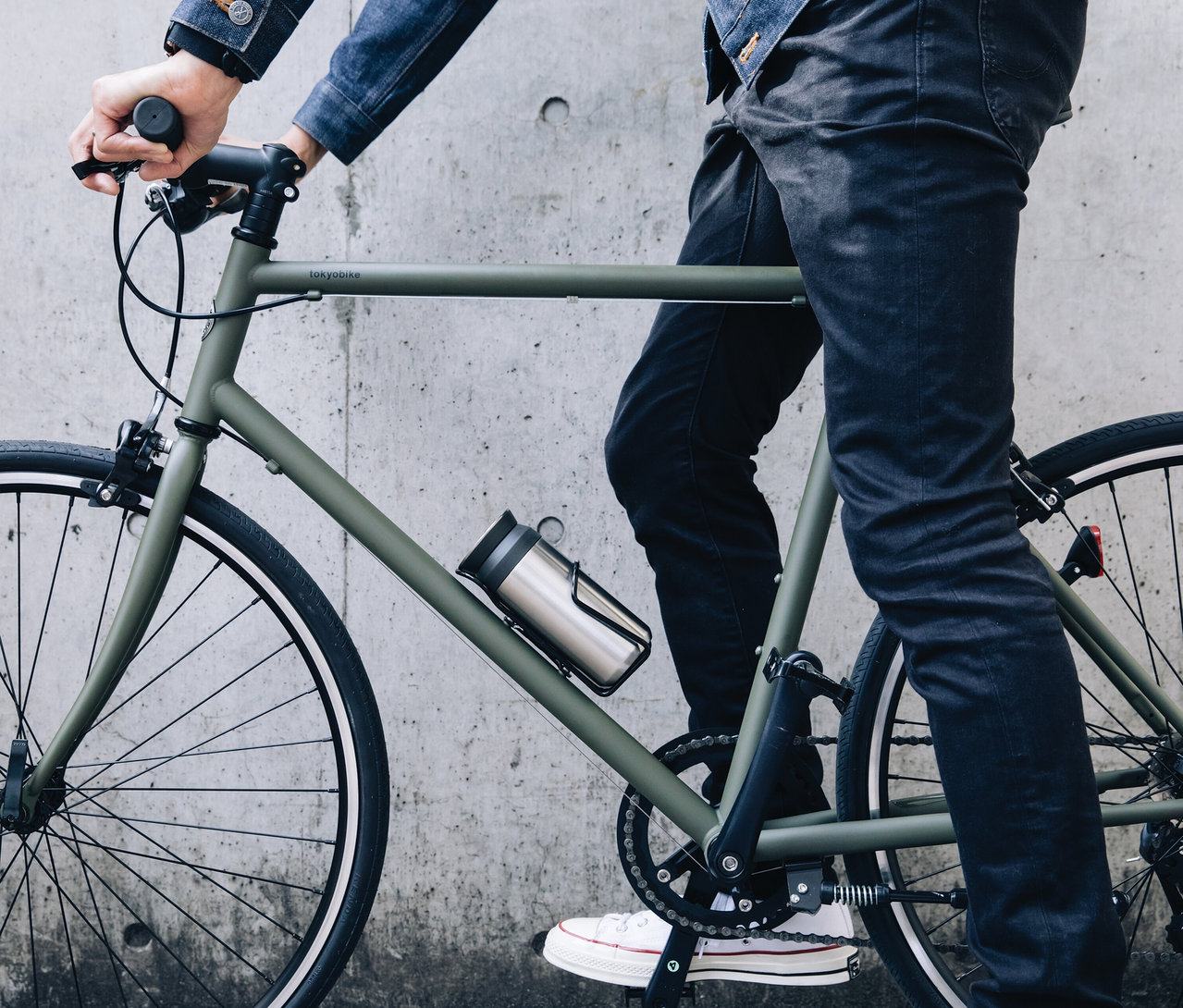 Mann fährt ein olivgrünes Fahrrad vor einer Betonwand. Eine silberne Wasserflasche befindet sich am Rahmen des Fahrrads.