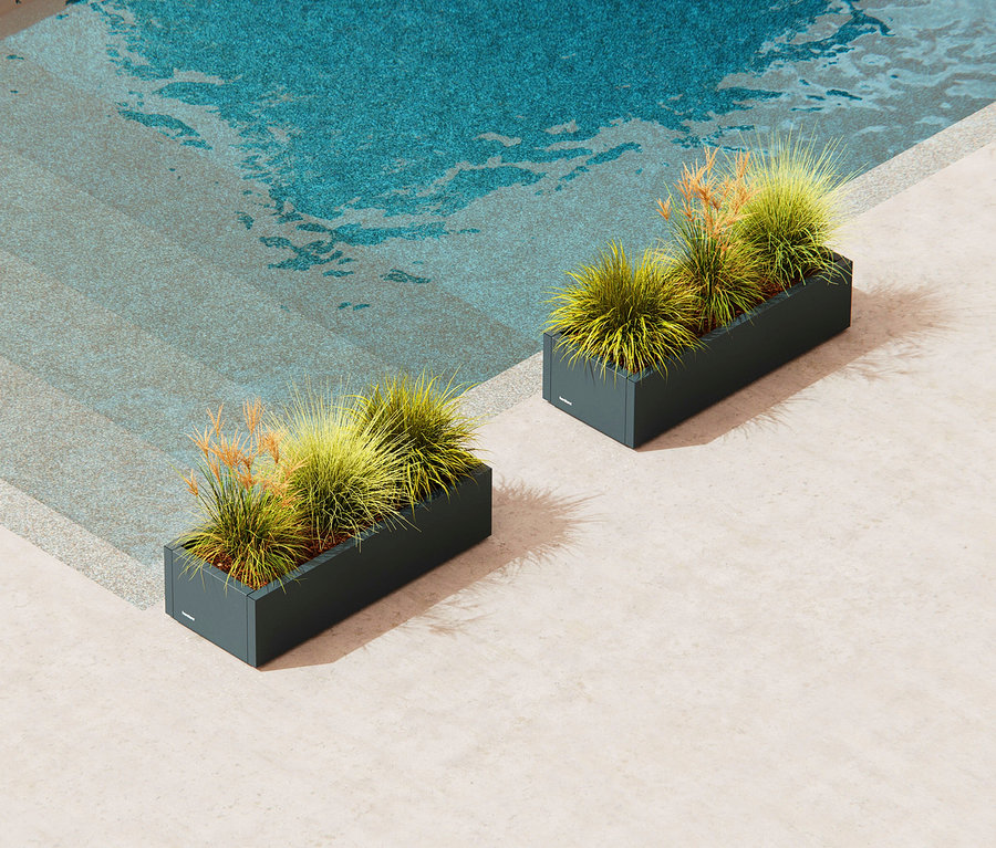 Zwei graue Pflanzkästen mit grünem und braunem Gras stehen auf einer Terrasse neben einem Schwimmbad.
