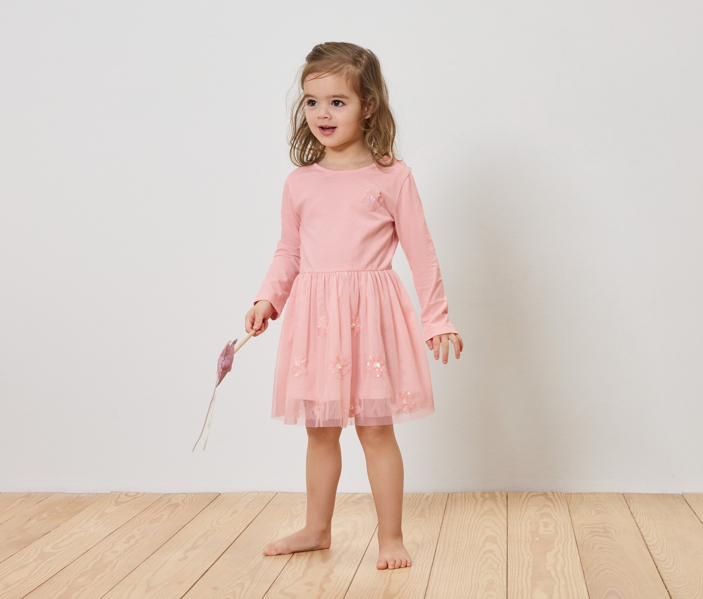 Mädchen mit blonden Haaren trägt ein rosa Kleid und hält einen lila Gegenstand in der Hand.