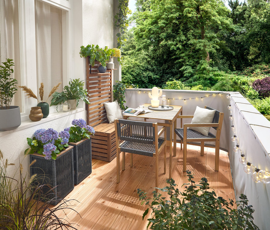 Einrichtung eines Balkons mit einem Balkon-Set mit Textilgeflecht, einem grauen Balkon Sicht- und Windschutz, einer LED-Outdoor-Lichterkette mit Netzadapter, einem Pflanztopf mit Textilgeflecht, einer LED-Outdoor-Akku-Tischleuchte in Kaktusform und 2 Outdoor-Kissen mit Leinen-Haptik.