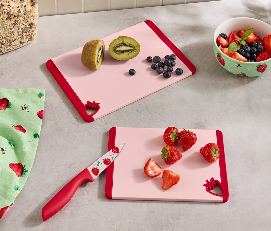 Auf einer Küchentheke liegen zwei rosa Schneidebretter mit rotem Rand, ein Behälter mit Müsli, eine Schüssel mit Beeren, ein Geschirrtuch und ein Messer.