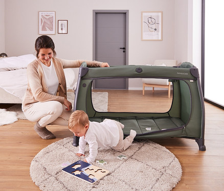 Eine Frau kniet neben einem olivgrünen Laufstall, während ein Baby davor auf einem Teppich mit einem Puzzle spielt.