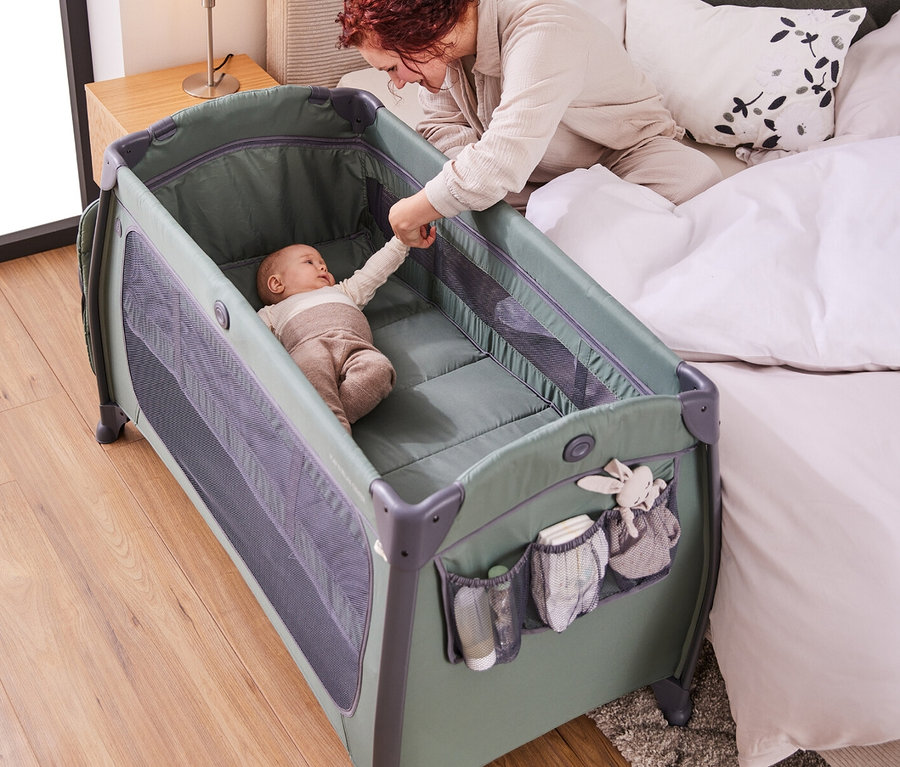 Eine Frau reicht einem Baby, das in einem olivgrünen Reisebett mit Netzseiten liegt, die Hand.