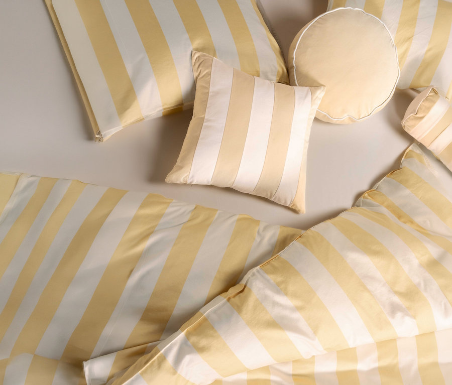 Draufsicht von Kissen und Bettwäsche mit gelben und weißen Streifen. Ein beiges Essenza Samt-Dekokissen »Nino« liegt mittig.