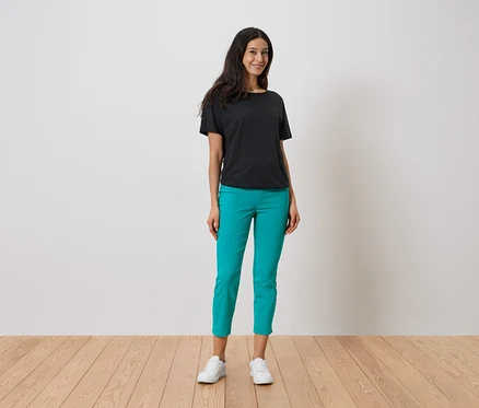 Frau steht und trägt ein schwarzes T-Shirt, eine türkisfarbene Hose und weiße Turnschuhe.