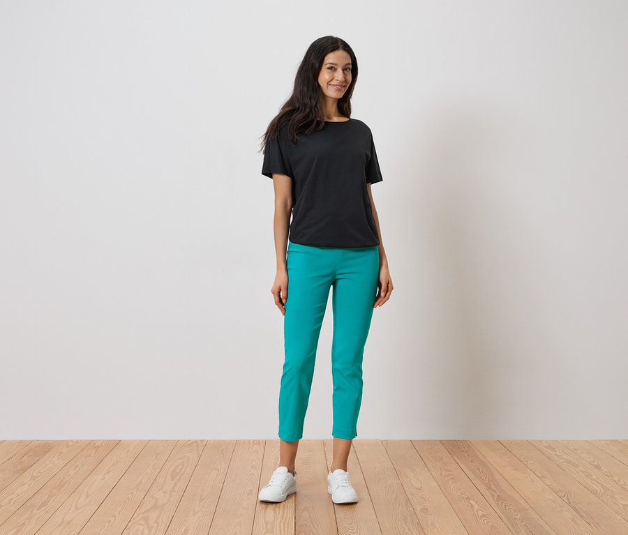 Frau steht und trägt ein schwarzes T-Shirt, eine türkisfarbene Hose und weiße Turnschuhe.