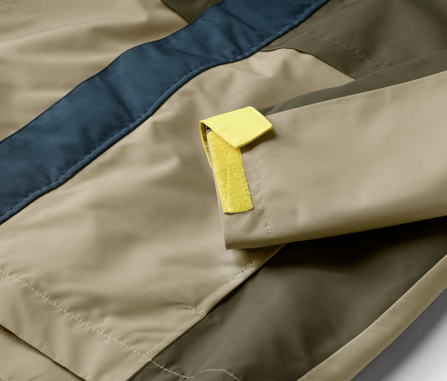 Nahaufnahme einer Jacke in Beige, Olivgrün und Dunkelblau mit gelbem Detail am Ärmel.