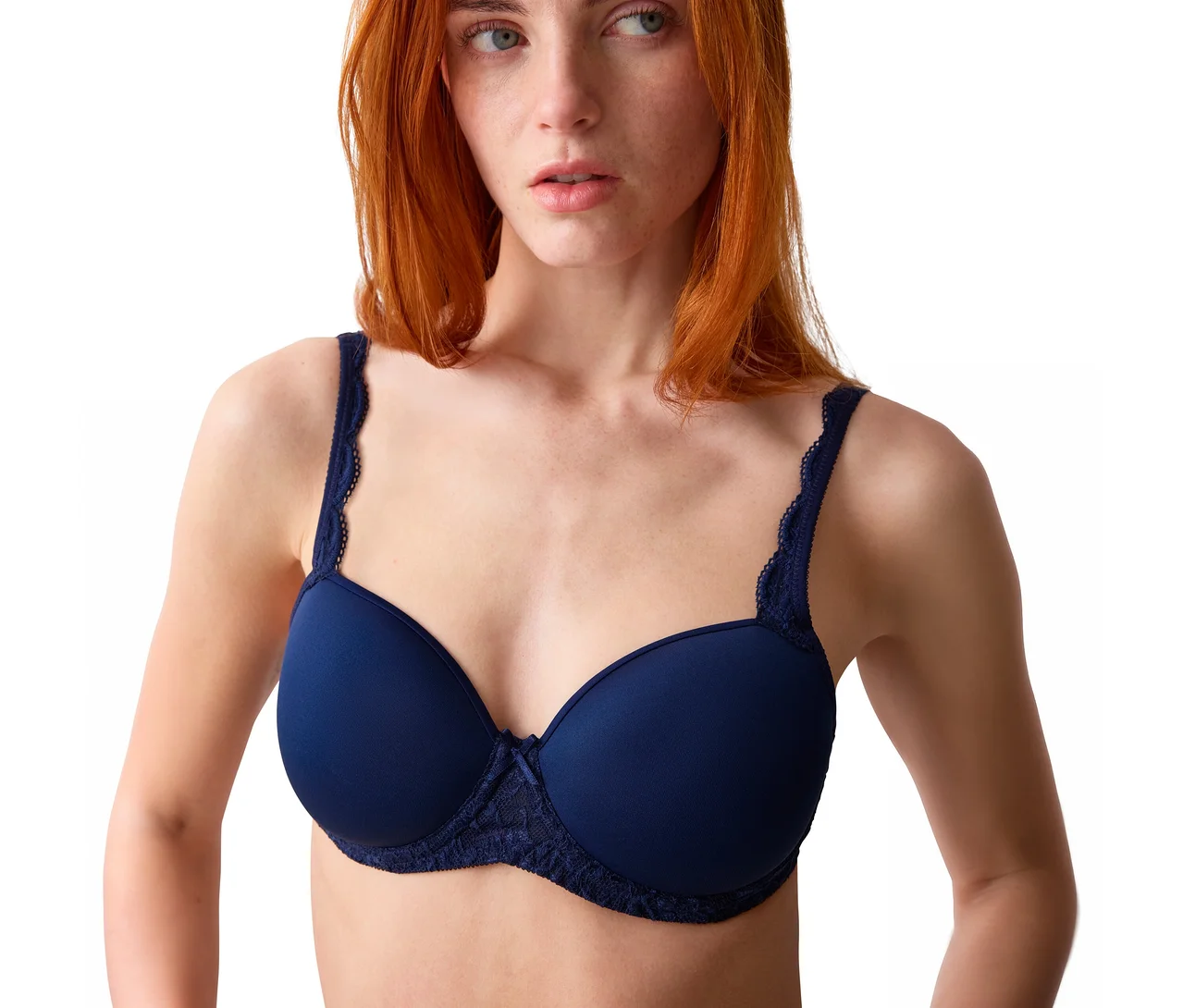 Eine rothaarige Frau mit Sommersprossen trägt einen marineblauen BH mit Spitzendetails an den Trägern und unter den Cups und blickt nach rechts vor weißem Hintergrund.
