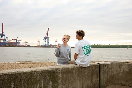 Frau lacht und hält einen Kaffeebecher in der Hand, Mann sitzt auf einer Mauer am Strand.