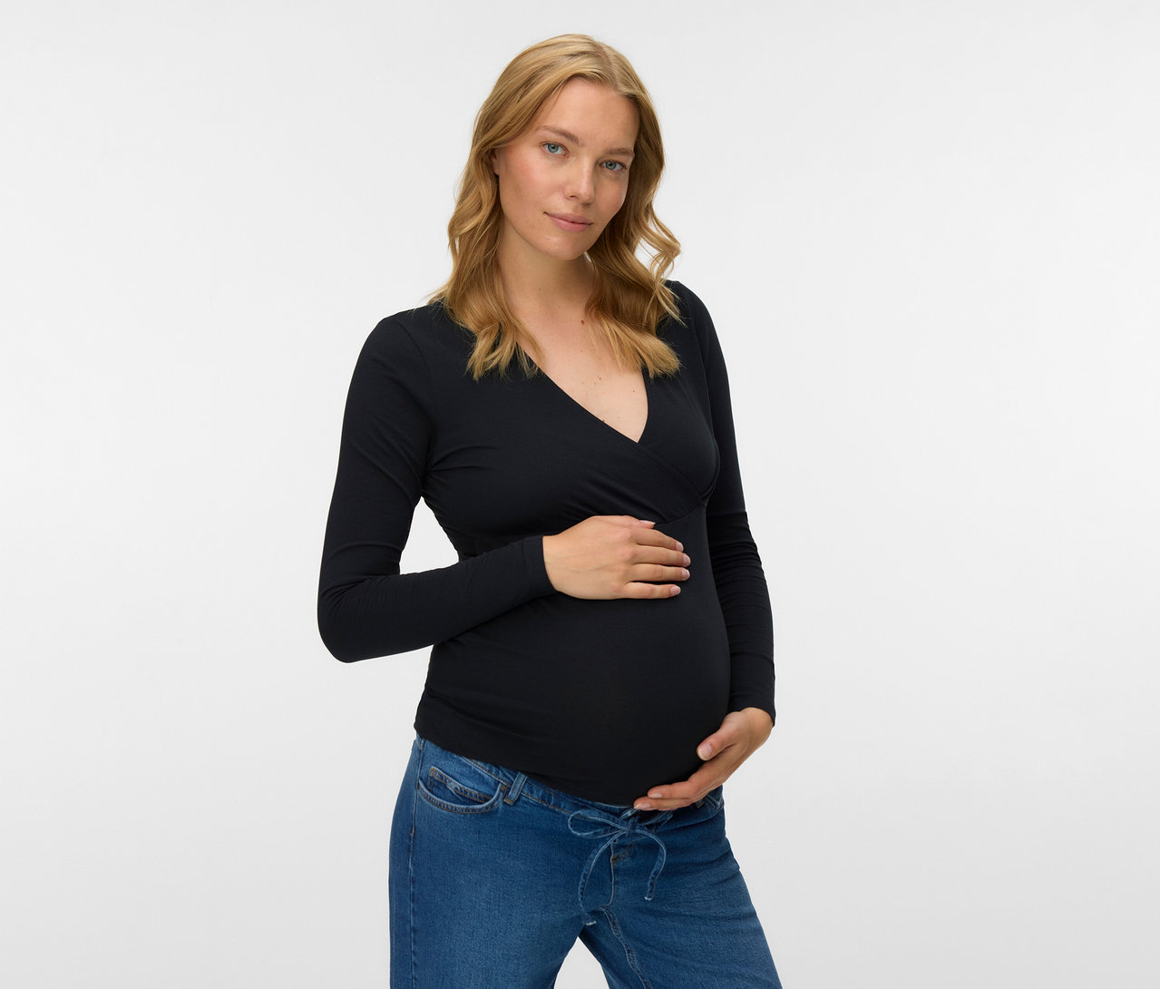 Schwangere Frau mit Jeans und schwarzem MAMALICIOUS 2er Pack Top »MLKERRIE« mit langen Ärmeln, die ihren Bauch hält.