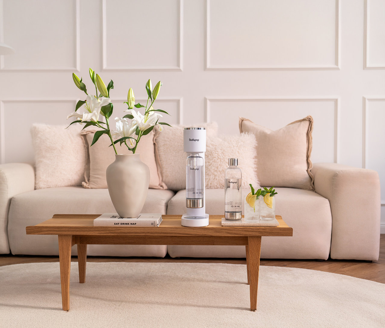 Auf einem Tisch stehen eine Vase mit Lilien, ein Buch, ein Sodapop Wassersprudler Cooper Pro in Weiß, eine Wasserflasche und zwei Gläser mit Zitronenscheiben.