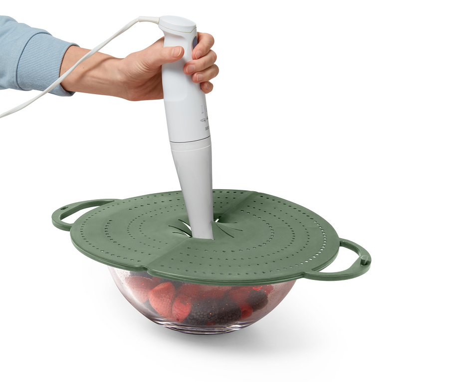 Eine Hand hält einen weißen Stabmixer über eine Glasschüssel mit Erdbeeren und einem grünen Deckel.