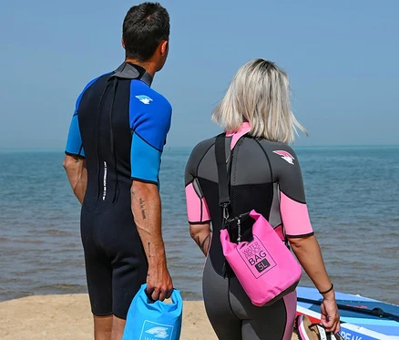 Zwei Personen in Neoprenanzügen stehen am Strand, blicken aufs Meer. Die Frau trägt eine pinke wasserdichte Tasche, der Mann eine blaue.