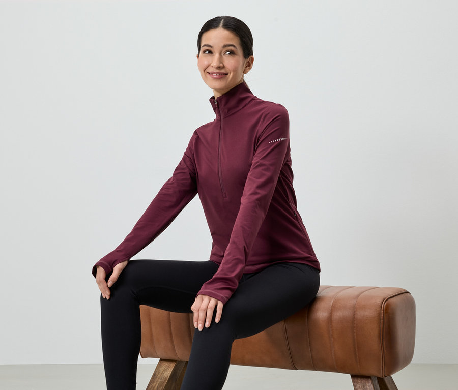 Eine Frau sitzt auf einem Hocker und trägt ein weinrotes, langärmeliges Oberteil und schwarze Leggings.