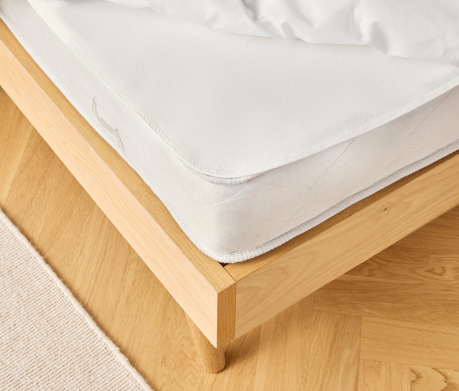 Detailansicht eines Bettes mit weißer Matratze und Bettwäsche auf einem hellen Holzboden, daneben ein beiger Teppich.