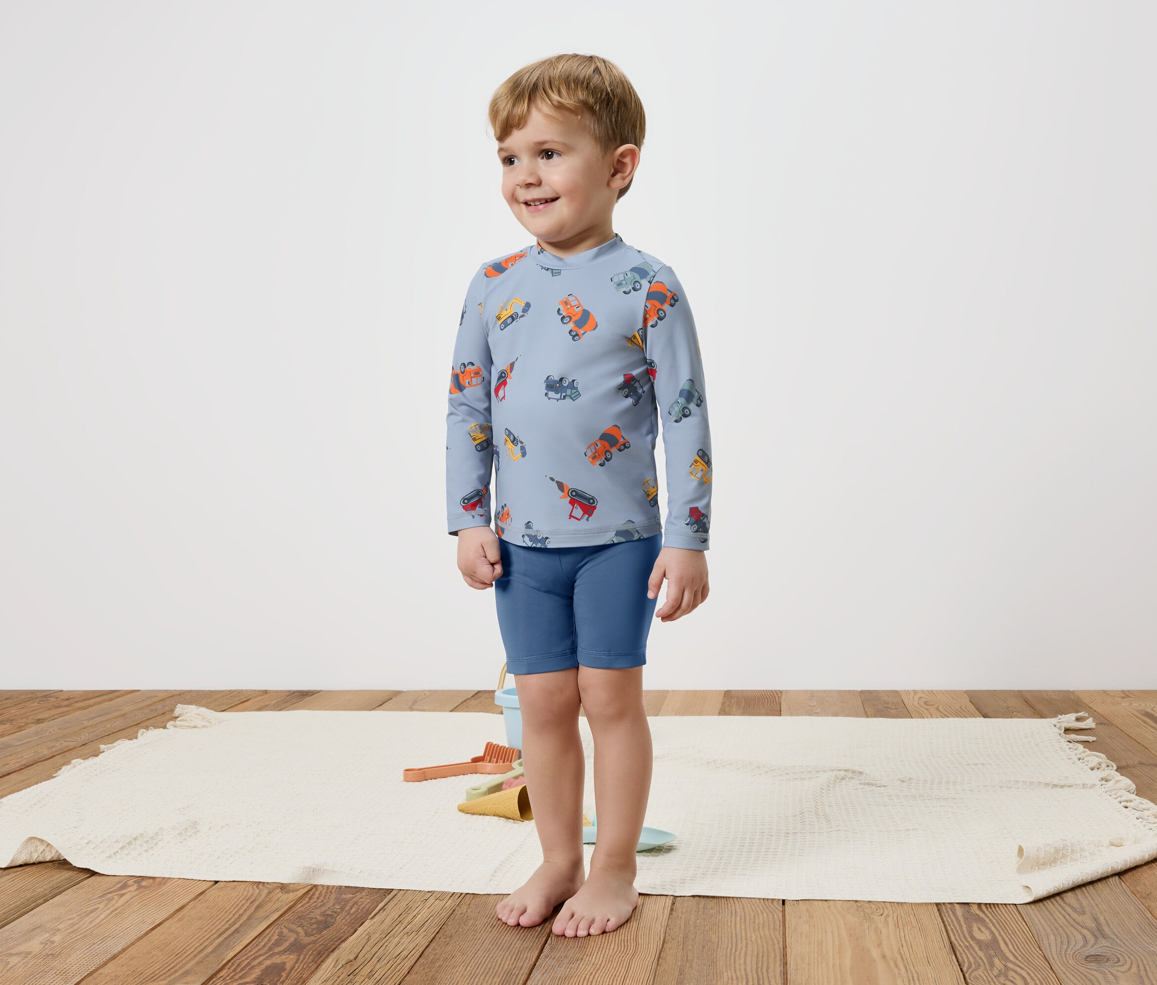 Ein kleiner Junge steht barfuß auf einem Teppich und trägt ein blaues, langärmliges Oberteil mit bunten Fahrzeugen und blaue Shorts.