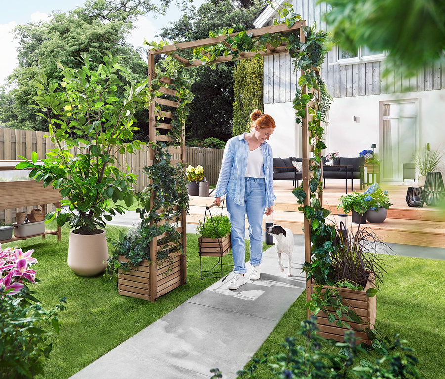 Eine Frau geht mit einem Hund durch einen Gartenbogen, der mit Pflanzen bewachsen ist. Sie trägt eine Pflanzbox.