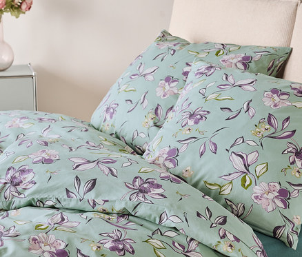 Bett mit hellgrüner Bettwäsche, die mit lila und weißen Blumen bedruckt ist. Zwei Kissen liegen auf dem Bett.