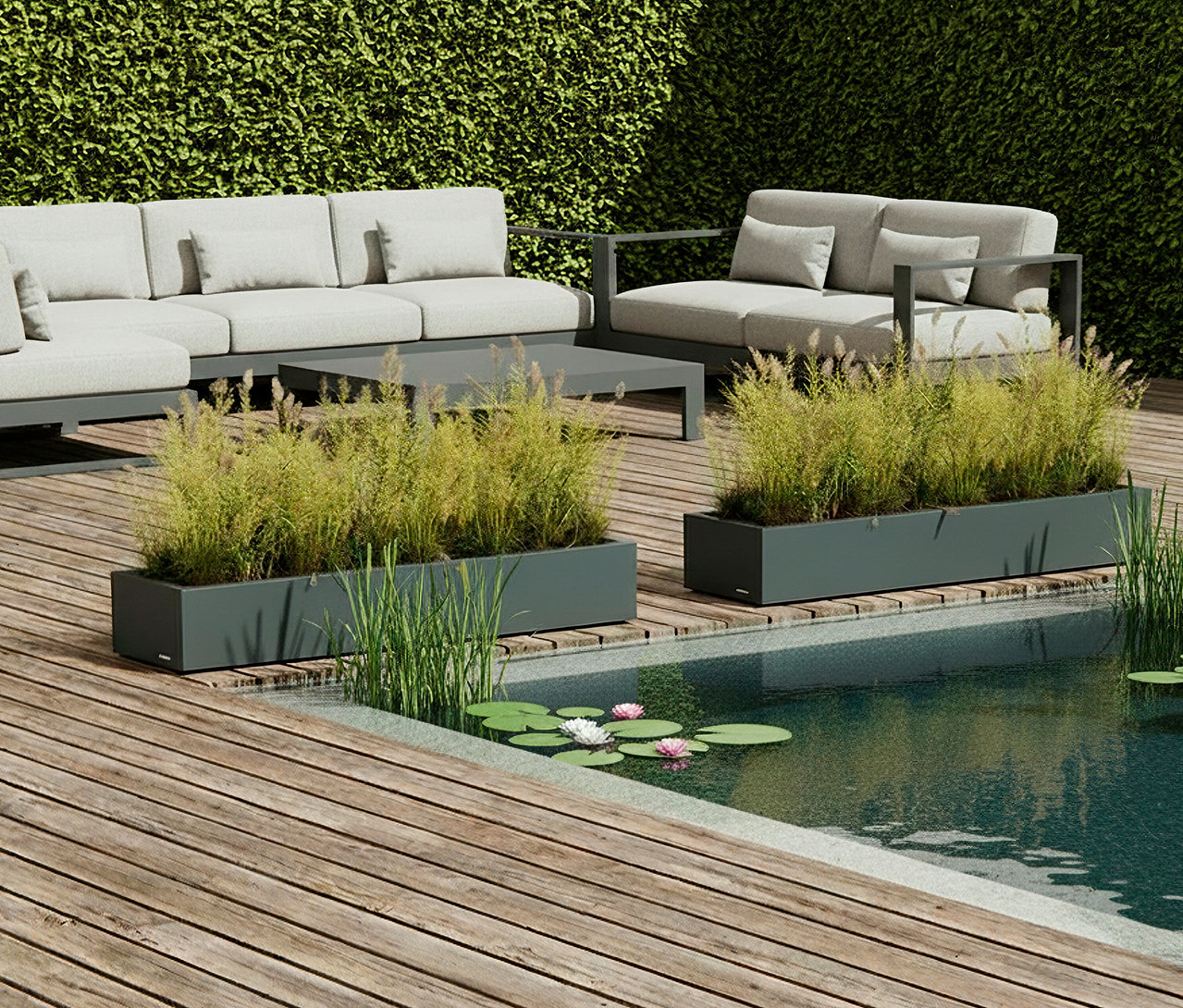 Auf einer Holzterrasse stehen graue Gartenmöbel und rechteckige Pflanzkästen mit Ziergräsern neben einem kleinen Teich mit Seerosen.