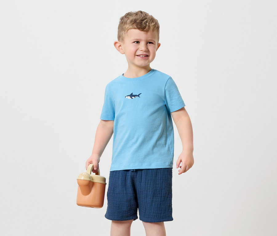 Ein kleiner Junge mit blonden Haaren trägt ein hellblaues T-Shirt mit einem Hai-Aufdruck und marineblaue Shorts. Er hält eine orangefarbene Gießkanne.