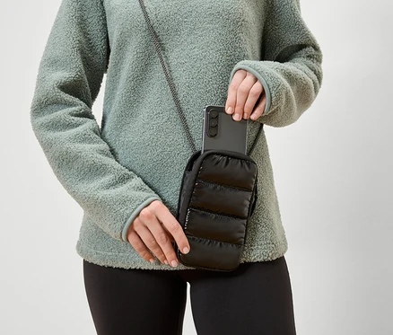 Frau legt ein Smartphone in eine schwarze, gesteppte Handytasche mit Kordel, die sie um den Hals trägt. Sie trägt ein grünes Fleeceoberteil.