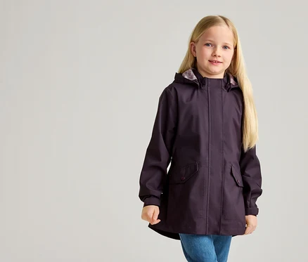Mädchen mit langen blonden Haaren trägt einen auberginefarbenen Regenmantel mit Kapuze und blaue Jeans.
