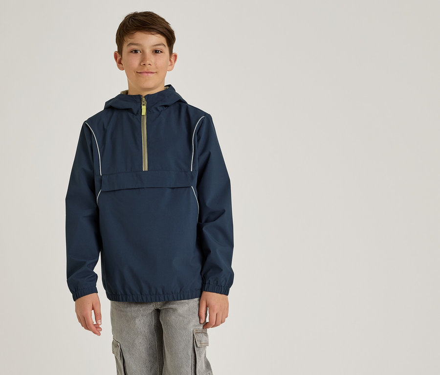 Ein Junge trägt eine marineblaue Anorakjacke mit Kapuze und graue Cargohosen vor einem schlichten Hintergrund.