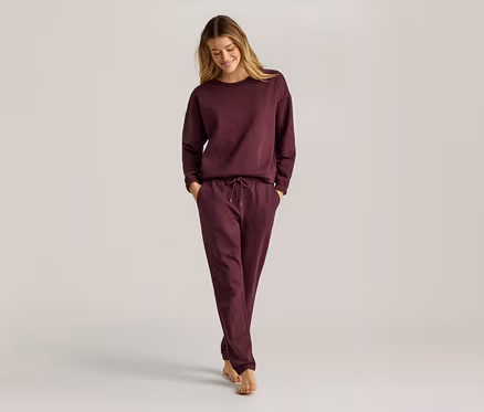 Frau mit langen blonden Haaren, die ein weinrotes, langärmeliges Sweatshirt und eine Jogginghose trägt und barfuß geht.