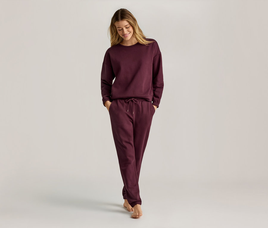 Frau mit langen blonden Haaren, die ein weinrotes, langärmeliges Sweatshirt und eine Jogginghose trägt und barfuß geht.