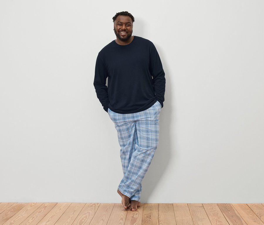 Ein dunkelhäutiger Mann mit Bart, der ein dunkelblaues langärmeliges Hemd und eine blau-weiß karierte Pyjamahose trägt, steht barfuß vor einer weißen Wand auf einem Holzboden.