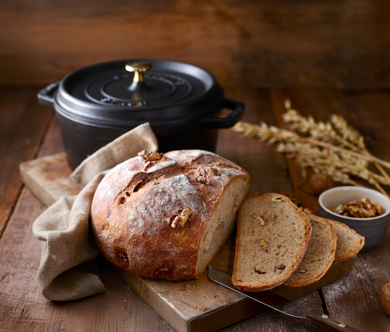 Ein Laib Nussbrot und Brotscheiben auf einem Schneidebrett mit einem schwarzen STAUB »Cocotte 24« Kochtopf im Hintergrund.