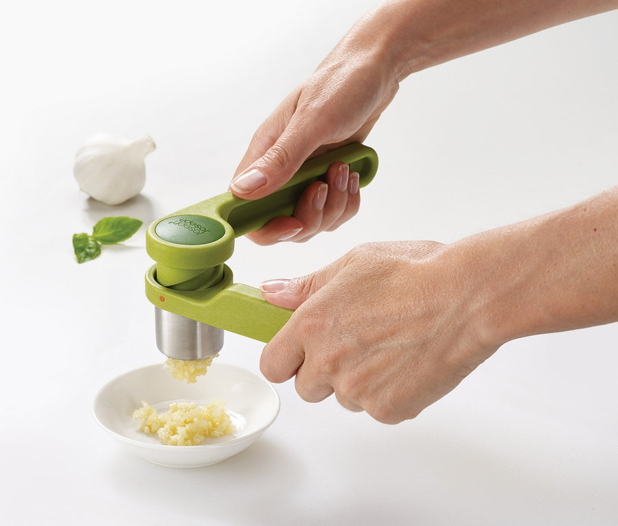 Eine Person presst Knoblauch mit einer grünen Knoblauchpresse in eine kleine Schale. Eine Knoblauchzehe und ein Basilikumblatt liegen daneben.