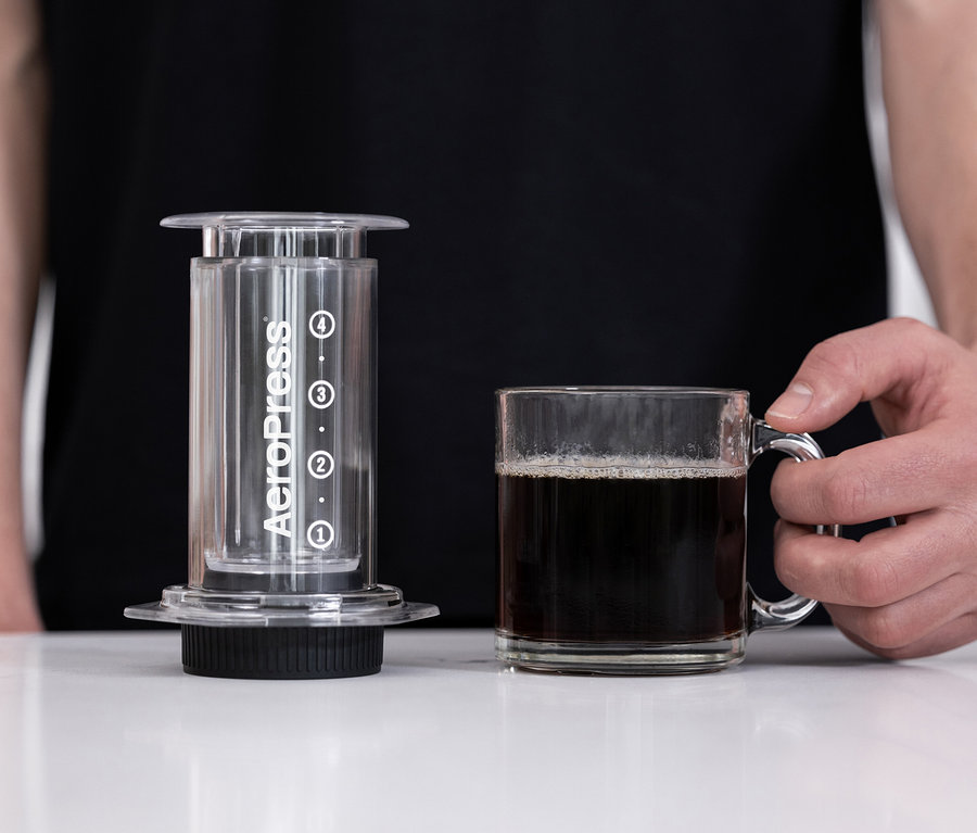 Eine Person hält eine Tasse Kaffee neben einer durchsichtigen AeroPress Kaffeemaschine.