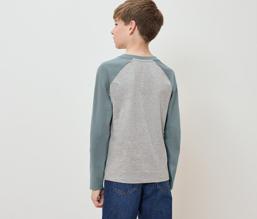 Junge steht mit dem Rücken zur Kamera, trägt ein graues T-Shirt mit langen grünen Ärmeln und blaue Jeans.