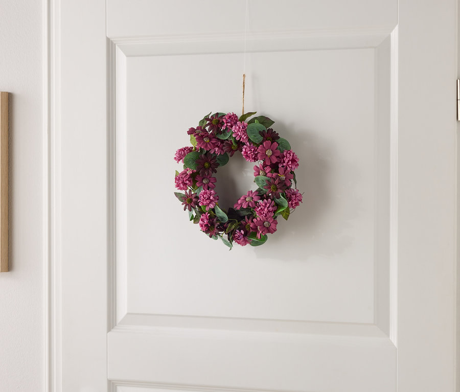 Kleiner Blumenkranz mit lila Blüten und grünen Blättern hängt an einer weißen Tür.
