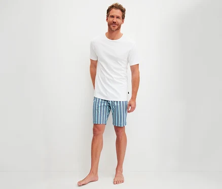 Lächelnder Mann trägt ein weißes T-Shirt und blau-grün gestreifte Shorts aus einem CALIDA Herren-Shorty-Pyjama-Set, barfuß vor weißem Hintergrund stehend.