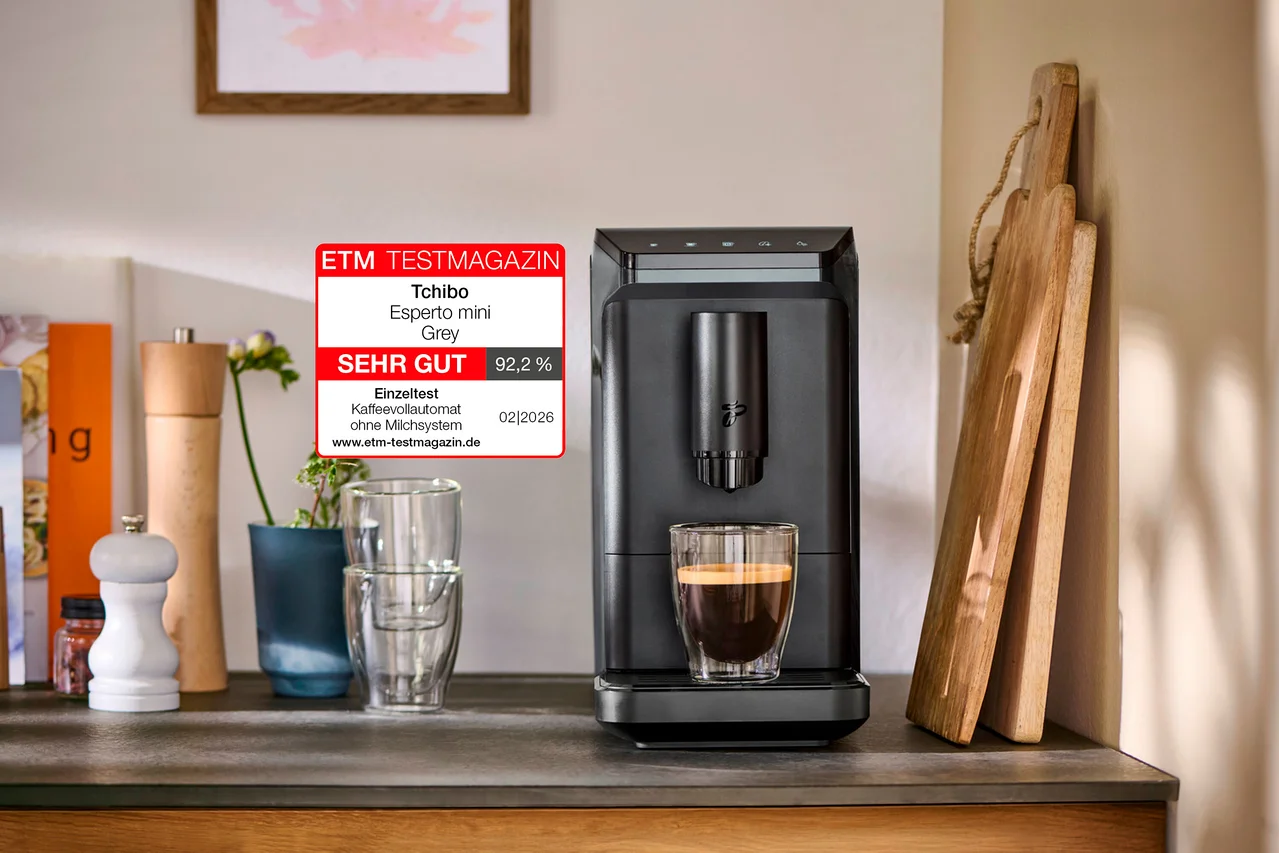 Schwarzer Tchibo Esperto mini Grey Kaffeevollautomat auf einer Arbeitsplatte, mit einer Tasse Espresso darunter. Ein ETM TESTMAGAZIN Aufkleber bewertet ihn mit SEHR GUT 92,2%. Daneben stehen Gewürzmühlen und Schneidebretter.
