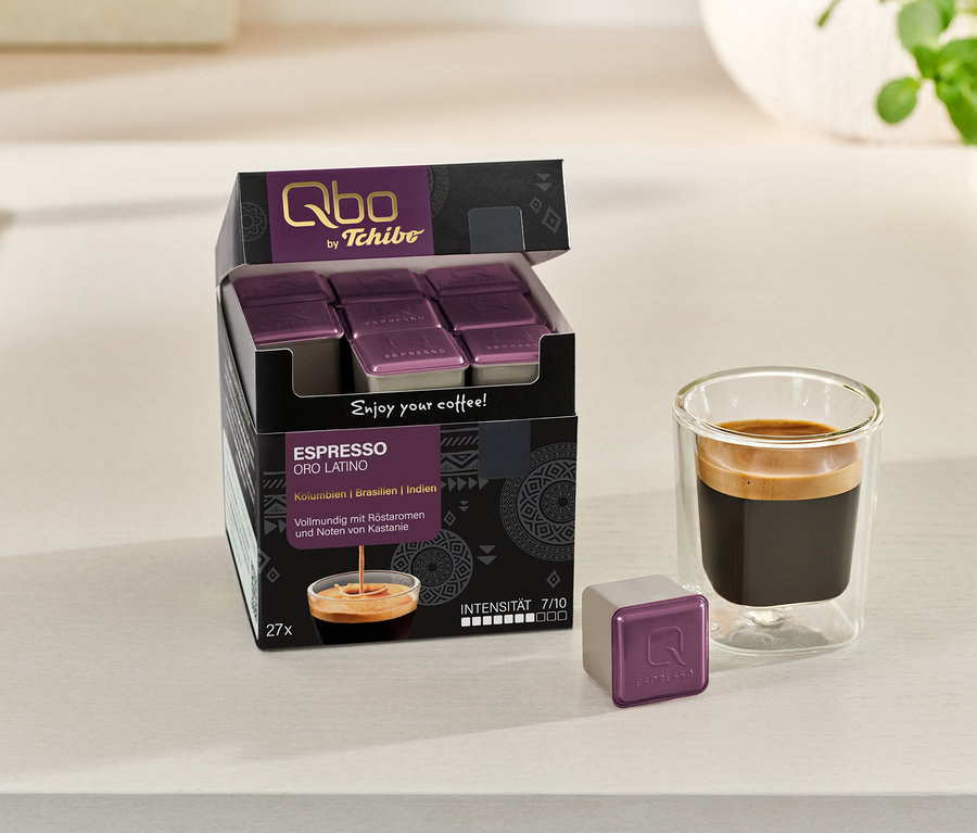 Ein geöffneter Karton Qbo Espresso ORO TOLIMA mit Kapseln steht neben einem Glas Espresso und einer einzelnen Kapsel.