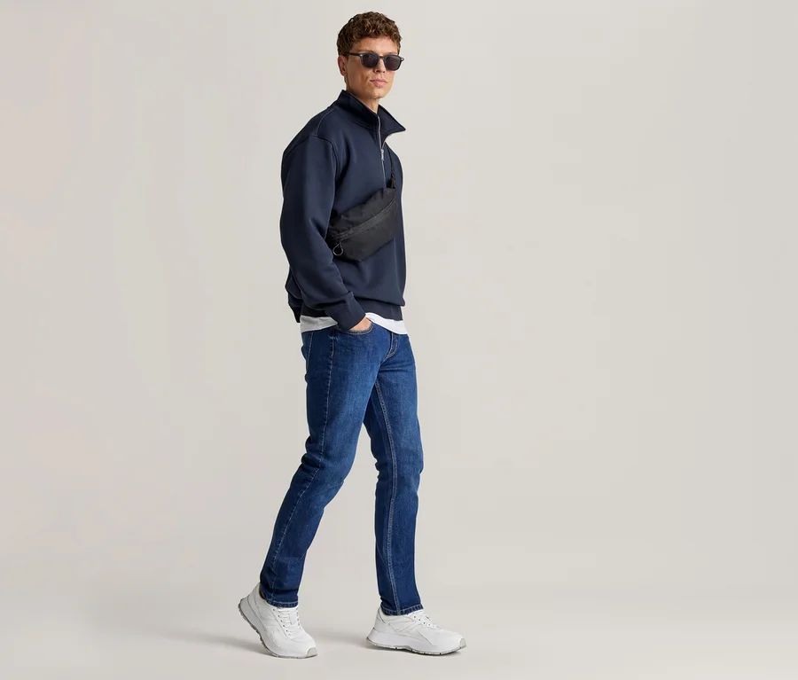 Mann geht, trägt Sonnenbrille, ein dunkelblaues Sweatshirt mit halbem Reißverschluss, blaue Jeans, eine schwarze Umhängetasche und weiße Sneaker.