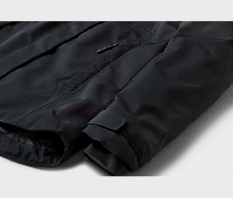 Nahaufnahme einer schwarzen Jacke mit einer Reißverschluss-Brusttasche, deren Zipper einen Schriftzug trägt, und einem verstellbaren Ärmelbündchen mit Klettverschluss.