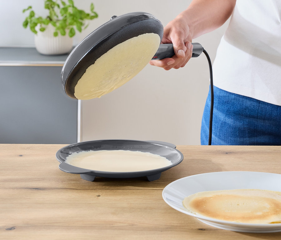 Eine Person neigt eine dunkelgraue elektrische Bratpfanne über einen Teller mit restlichem Teig. Ein fertiger Crêpe liegt auf einem weißen Teller auf einer Holzablage.