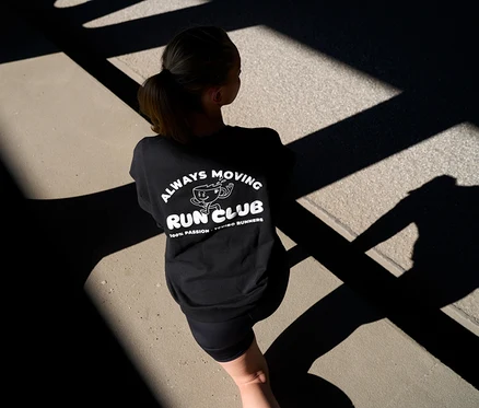 Frau von hinten in schwarzem Sweatshirt mit „Always Moving Run Club“-Aufdruck und kurzen anthrazitfarbenen Sportleggings »ActivePower« auf einem Weg mit Schatten.