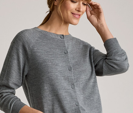 Lächelnde Frau mit hellbraunem Haar, trägt einen grauen Cardigan über einem weißen Langarmshirt und blaue Jeans. Ihre linke Hand steckt in der Hosentasche, die rechte berührt ihre Schläfe.