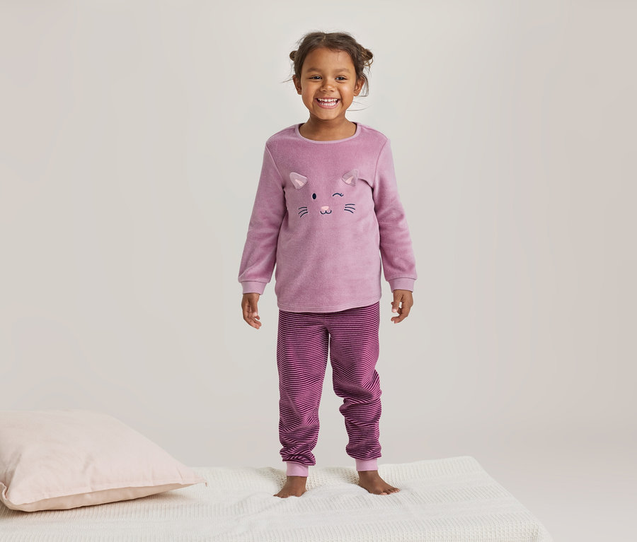 Lächelndes kleines Mädchen in einem rosa Pyjama mit Katzenmotiv und gestreiften Hosen steht auf einer weißen Decke neben einem rosa Kissen.