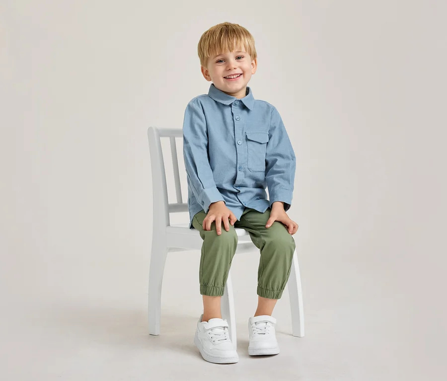 Lächelnder kleiner Junge mit blonden Haaren sitzt auf einem weißen Stuhl und trägt ein blaues Hemd, olivgrüne Hose und weiße Turnschuhe.